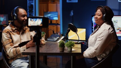 African american influencer using professional camera to record podcast with guest, talking and having fun together. Happy host discussing with woman live in apartment studio for internet show