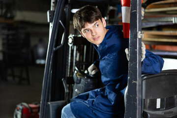 Focused young workman in blue work coat operating lift truck in industrial warehouse setting, looking over shoulder while maneuvering vehicle