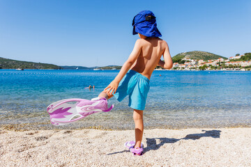 Back view cute little tanned boy kid in swimsuit sunscreen cap and fin funny walking dive in clear...