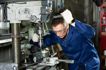 Confident skilled operator in blue workwear and protective glasses, working on drilling machine in metalworking shop, performing holes in flat steel part