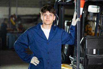 Smiling young male forklift operator in blue work coat and gloves standing with hand akimbo beside...