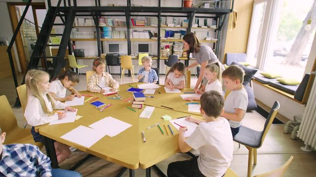 Elementary school students sit at a round table in an art class, and a young, modern teacher in glasses helps them with their classwork