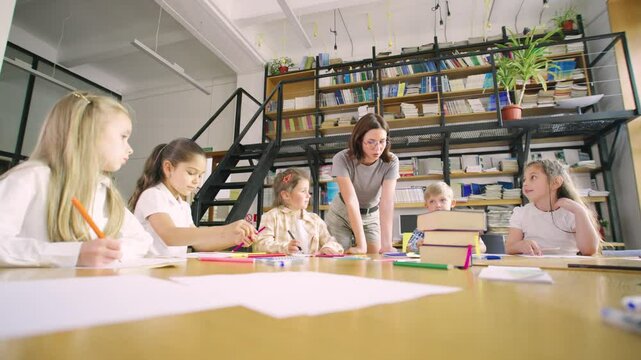 Elementary school students sit at a round table in an art class, and a young, modern teacher in glasses helps them with their classwork