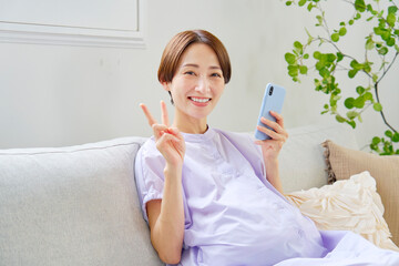 自宅でスマートフォンを持ってピースサインをする高齢妊娠をした妊婦