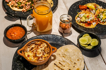 Queso Fundido with Chorizo ( Chorizo with melted cheese) and flour tortillas, mango soda, pastor tacos with red sauce and lemons in a white wooden background