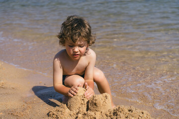 A young boy with curly brown hair is seated on the sandy shore of a beach, building sandcastles with his hands. The waves are gently lapping at the shore behind him, creating a peaceful scene