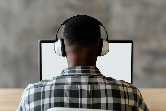 A young african american man sitting in front of a white laptop screen in his room back to camera