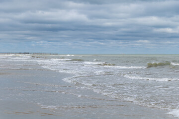 A wide shot of the water on a sandy beach with waves crashing against the shore and a pier visible in the distance, under a cloudy sky with overcast weather