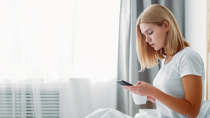 Media morning. Mobile routine. Modern life. Focused confident woman using phone drinking tea browsing surfing internet in light room with free space.