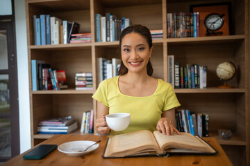 Young woman reading book and drink hot coffee in library at home, smiling, laughing, enjoying bookworms hobby, home leisure time