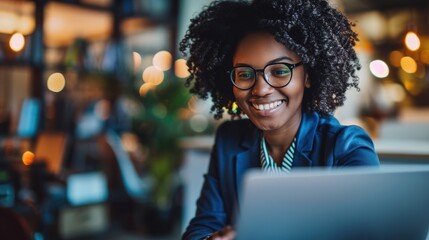 Smiling woman with glasses and curly hair working on a laptop in a modern, warmly lit office environment.