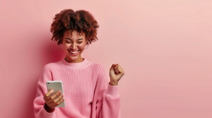 African American woman smiling at her phone, celebrating with a raised fist while holding her phone, concept of winning, solid pink background.