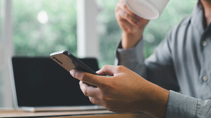 Close up of casual man or freelancer working on laptop and holding mobile smart phone with cup of coffee on table in coffee shop or cafe, working from cafe, freelance working concept.
