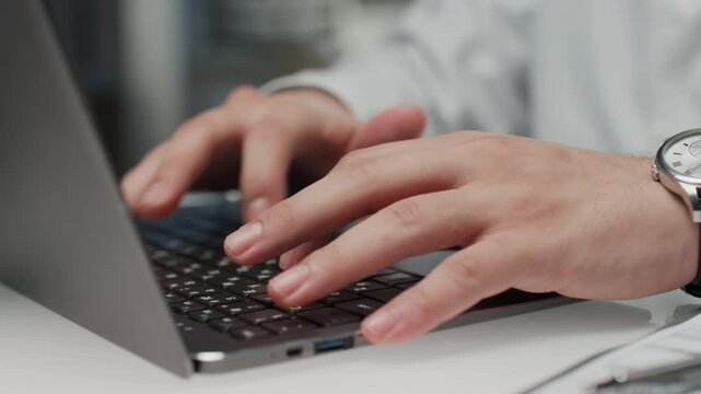 Panning closeup of hands with watch of unrecognizable medical worker in lab coat filling in online treatment plan while typing on wireless laptop computer at workplace in hospital