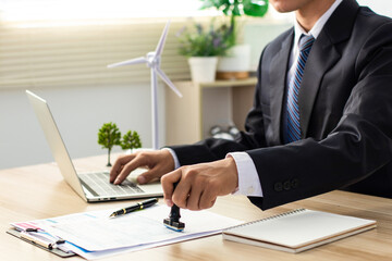A businessman in a suit stamps his approval on a document, with a miniature wind turbine visible on...
