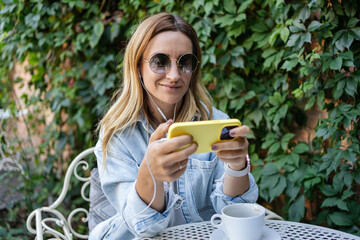  woman browsing on phone while wearing earphones and listening to music while sitting at outdoor cafe. Freelancer working remote on restaurant. 