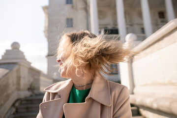 A woman with long hair is standing on a set of stairs. She is wearing a green shirt and a tan coat.