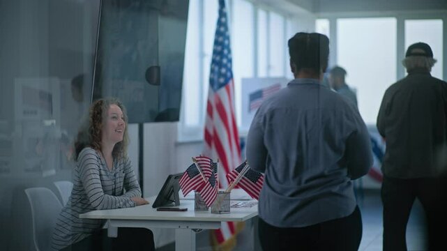 African American woman approaches registration table at polling station. Female polling worker talks to woman and uses tablet computer. Diverse people vote in voting booths. US presidential elections.