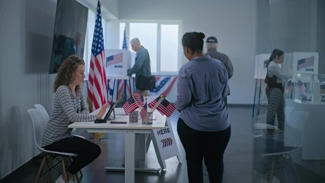 African American woman comes to registration table at polling station. Female polling worker talks to woman and uses digital tablet. Multiethnic US citizens vote in voting booths. Election Day in USA.