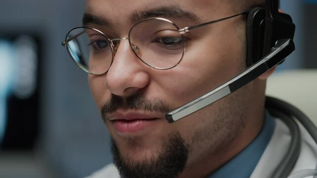 Closeup of positive male Biracial doctor with moustache and beard, wearing glasses and on-ear call center set, having conversation with patient and answering questions in modern hospital