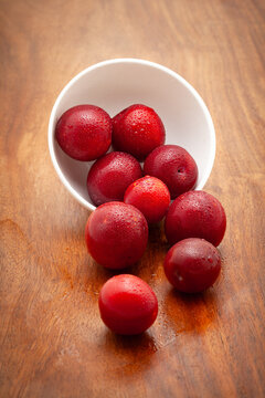 Front view of fresh Cherry Plum (Prunus cerasifera) fruits spilled out from a white ceramic bowl, on a wooden background.