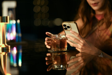 Charming young woman hand holding a glass of whisky using smartphone at bar counter