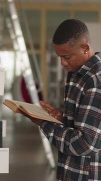 Dark-skinned man peruses book near bookshelf. African American student studies book fluently before picking up for home reading. Guy reads novel