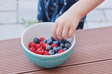 Child hand pick berries from bowl. Blueberries and raspberries. Family and lifestyle. Backyard in summer and nature, terrace. 