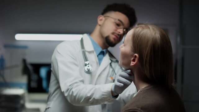 Medium shot of young bearded Biracial doctor wearing glasses, scrubs and stethoscope around neck examining sitting female patient, palpating lymph nodes on neck during annual checkup in clinic