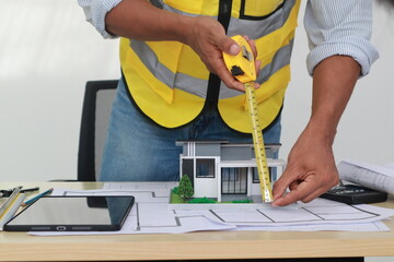 An engineer meticulously draws house plans on a workbench, surrounded by tools and blueprints,...