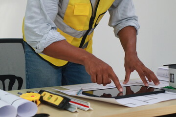 An engineer meticulously draws house plans on a workbench, surrounded by tools and blueprints,...
