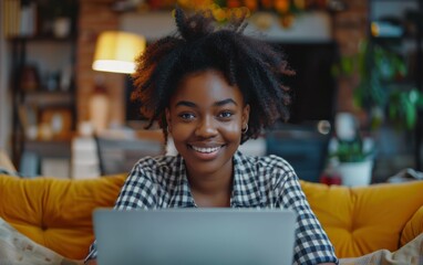 Cheerful black woman with curly hair working at home on laptop, smiling. Sitting by window with green plant in foreground, garden view outside