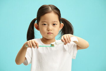 Japanese child girl squeeze toothpaste from tube on toothbrush and look to camera, standing over blue isolated background. 