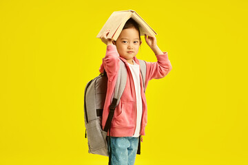 Cute chinese elementary school girl holding book above head and having backpack on shoulders, standing on yellow isolated background. Asian ethnicity six year old kid preparing to get school. 