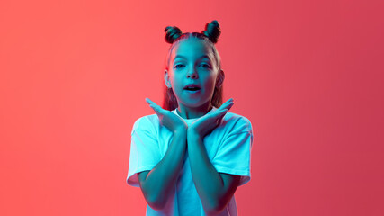 Portrait of young girl in white t-shirt, with hair styled in two buns posing against vibrant pink background in neon light. Concept of childhood, human emotions, casual fashion