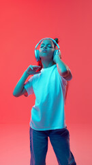 Portrait of young active girl in casual clothes, listening to music in headphone witch involved face against pink background in neon light. Concept of childhood, human emotions, casual fashion
