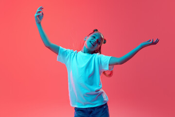 Portrait of young girl in white t-shirt and jeans spreading hands in positive expression against pink background in neon light. Concept of childhood, human emotions, casual fashion
