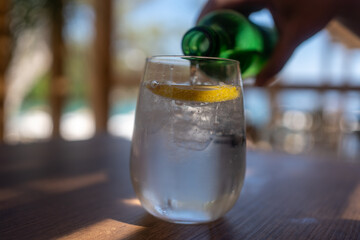 Image of pouring water from a water bottle into a glass at beach bar. Drink and refreshment concept - glass of sparkling water with lemon slices on table.