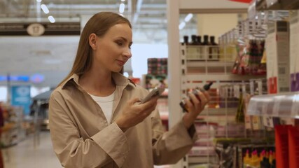 A woman in a beige shirt scans a package of cosmetics in a retail store. She is using a smartphone to scan the product, likely to check prices or read product information