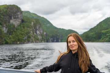 Female tourist with brown hair in a black hoodie smiles on a catamaran with fjord view in Norway.