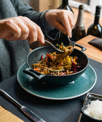 A person is twirling pasta with a fork in a black bowl, garnished with vegetables. The setting is a cozy restaurant with natural light, perfect for culinary and dining content.