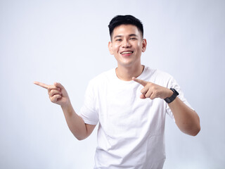 A young Asian man, wearing a white shirt and black watch, smiles widely while pointing both index...