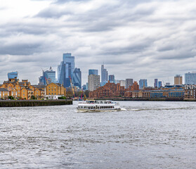 Obraz premium Ferry or tourist boat in the foreground sailing on the River Thames under a cloudy sky and old buildings on the river walk and skyscrapers on the horizon.