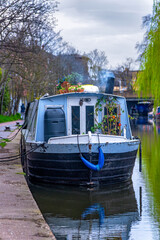 Naklejka premium Beautiful narrow blue wooden boat or houseboat docked and moored in the residential district of Maida Vale in London, with flower pots on its roof and smoke coming from its chimney.