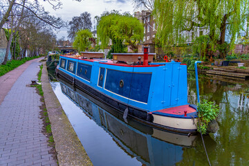 Narrow blue wooden narrowboats or houseboats docked and moored on the river in the residential district of Maida Vale. London with the vision of the promenade and the typical English houses.