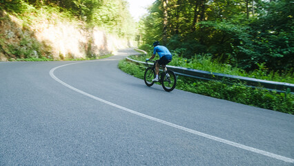 Obraz premium Male road racing cyclist in a blue sports jersey, with helmet and glasses, sprinting up a hill through the forest.