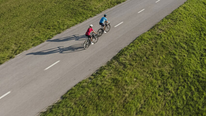 Side view of a Caucasian man and woman, in cycling wear, training for a race, riding road bikes in the middle of the paved road, aerial tracking drone shot.