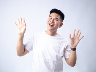 A young Asian man, wearing a white shirt and black watch, has a surprised and exaggerated expression with his mouth open and hands spread wide. The plain white background highlights