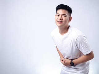 A young Asian man, wearing a white shirt and black watch, makes a dramatic expression of pain or discomfort while clutching his stomach. The background is plain white