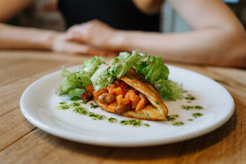 Close-up of delicious vegetable taco garnished with fresh lettuce and herbs, served on white plate with person in background. Gourmet Vegetarian Dish with Fresh Salad on White Plate at Restaurant. 
